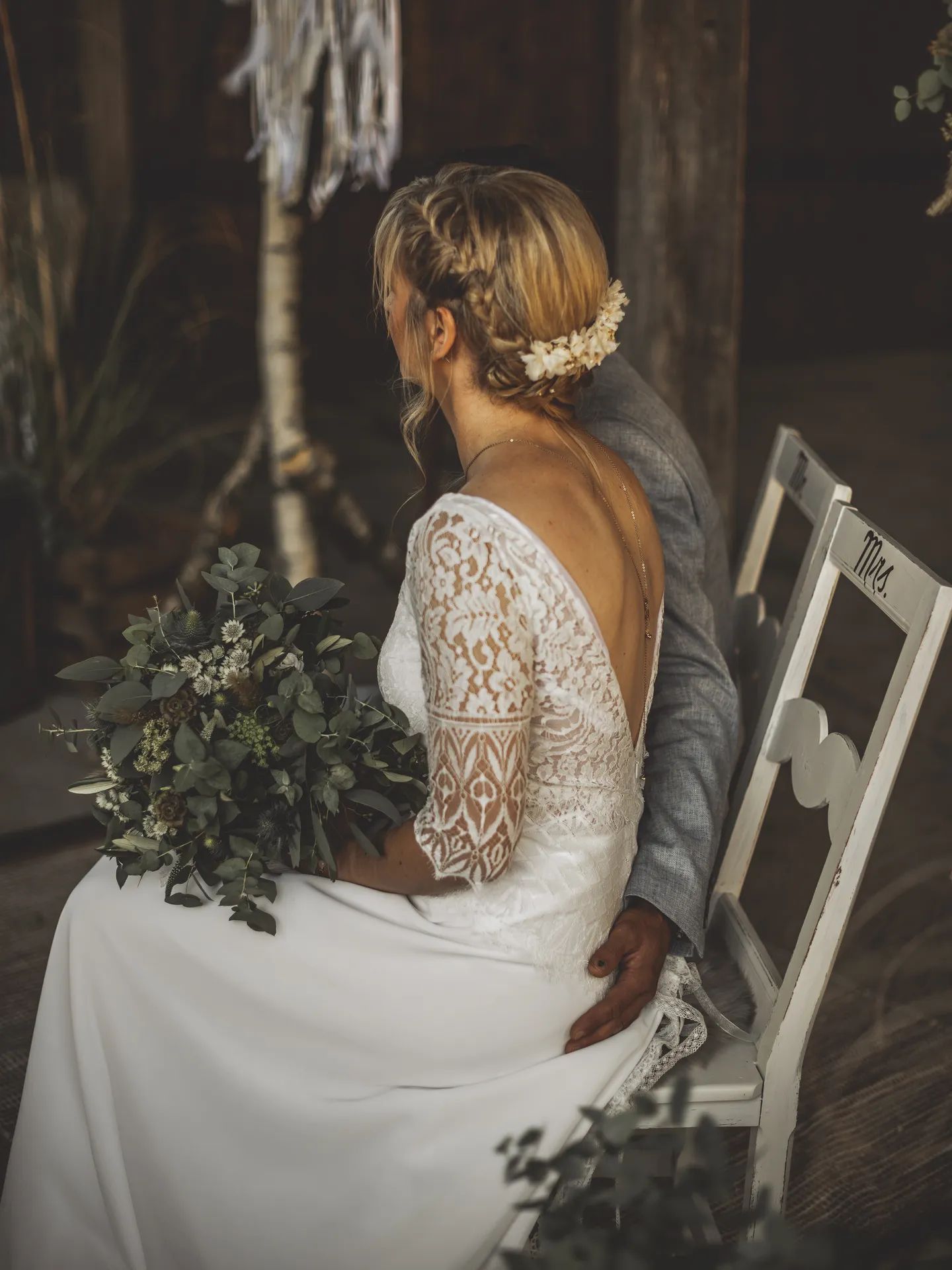 Mariée de dos, fleurs dans les cheveux et bouquet de verdure sur les genoux